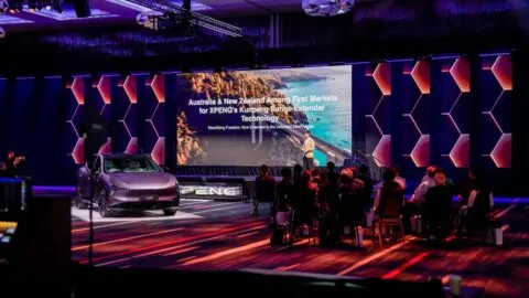An Xpeng L03 SUV beside a large presentation screen at an event with seated attendees.